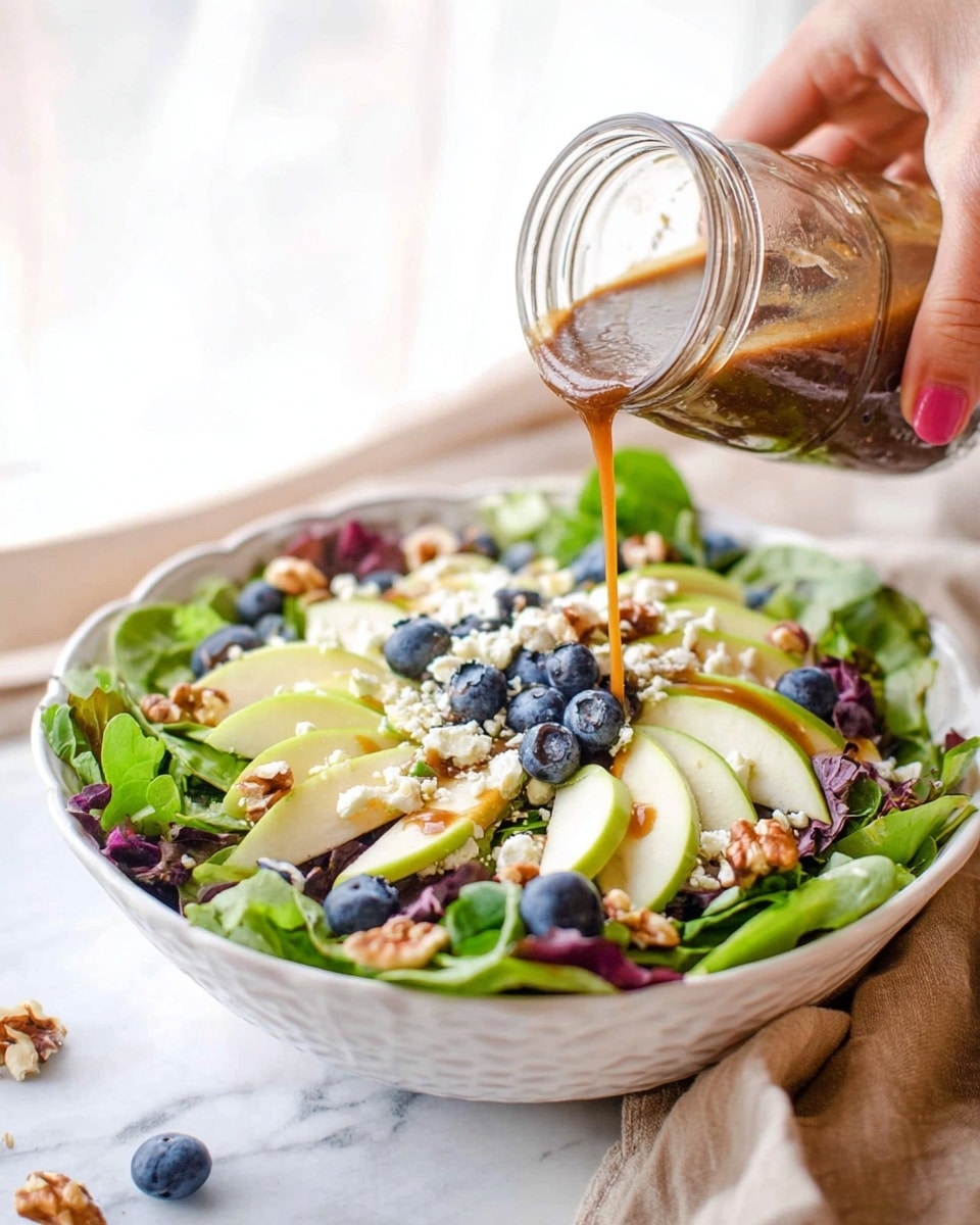 A white bowl filled with a fresh salad with multiple layers: the bottom layer is mixed green leaves with a mix of light and dark green, and dark purple colors; on top of the leaves are thin slices of light yellow apples, scattered blueberries in deep blue, creamy white cheese crumbles, and small light brown walnut pieces. A woman's hand is seen pouring a brown dressing over the salad from a glass jar. The scene is set on a white marbled surface with a soft natural light coming from behind. photo taken with an iphone --ar 4:5 --v 7