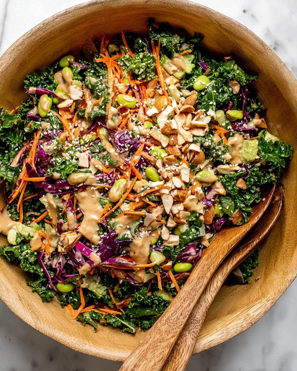 A large light wooden bowl holds a colorful salad with many layers. The bottom layer is dark green, curly kale leaves mixed with thin purple cabbage strips. Scattered on top are sliced red onions, orange shredded carrots, and pieces of light green edamame beans. There are also chopped almonds giving crunchy texture throughout. Creamy beige dressing is spread unevenly over the salad, and white sesame seeds are sprinkled on top for extra detail. Two wooden salad spoons are placed inside the bowl on the right side, partially covered by the salad. The bowl rests on a white marbled surface. photo taken with an iphone --ar 4:5 --v 7