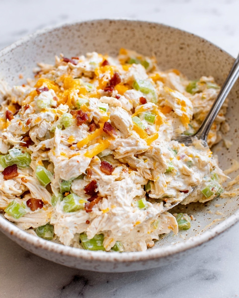 The image shows a close-up of a white speckled bowl filled with a creamy mixture. The mixture has several layers: a base of white creamy sauce, shredded pieces of light beige chicken, small chunks of light green celery, and bits of orange cheddar cheese mixed throughout. Tiny reddish-brown pieces, likely bacon, are scattered in the mix, adding contrast. The texture looks soft and thick with visible bits of ingredients. A silver fork is partially dipped into the mixture on the right side, with some sauce clinging to it. The bowl sits on a white marbled surface. Photo taken with an iphone --ar 4:5 --v 7