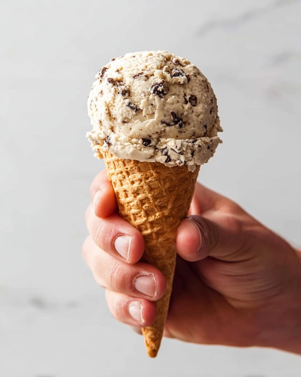 A close-up of a single scoop of light beige ice cream with small dark chocolate chips mixed throughout, sitting atop a waffle cone with a golden-brown color and a textured grid pattern. The ice cream scoop is round and slightly rough on the surface, showing the creamy texture and scattered chocolate pieces. The cone is held firmly by a man's hand with visible fingers and nails, against a clean white marbled background. photo taken with an iphone --ar 4:5 --v 7