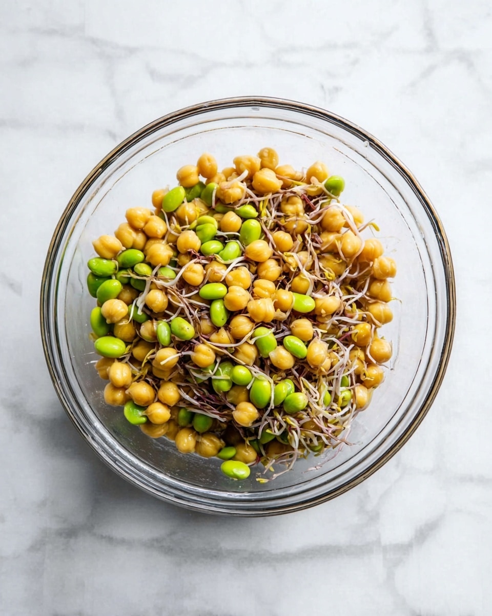 The image shows a clear glass bowl filled with a mixture of yellow chickpeas, bright green edamame beans, and thin brown bean sprouts. The ingredients are mixed evenly, creating a colorful and fresh-looking combination. The bowl is placed on a white marbled surface. photo taken with an iphone --ar 4:5 --v 7